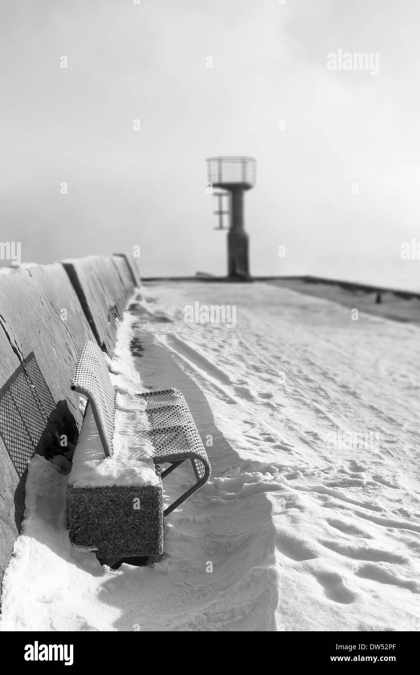 Blue metal bench on snowy mole and small beacon or lighthouse at the end of the pier Stock Photo
