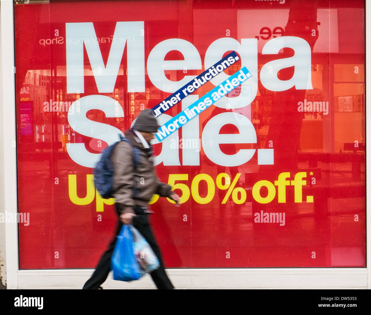 A motion blurred shopper carrying bags walking past a retail shop window display with a large red sale sign advertising discount Stock Photo