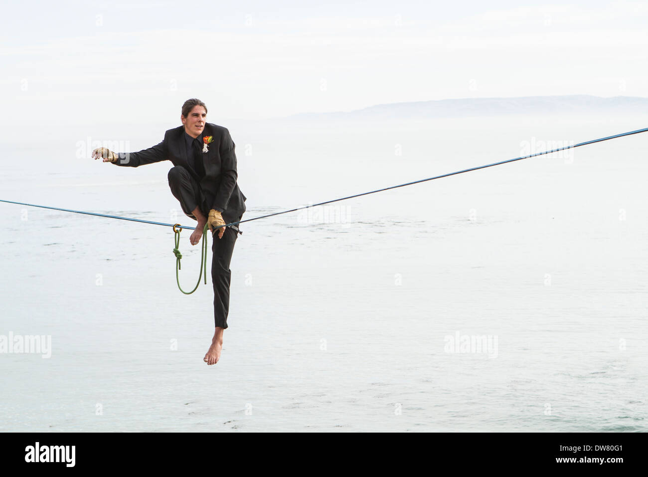 A groom in a suit goes to stand on a slackline above water to walk to his bride to be. Stock Photo