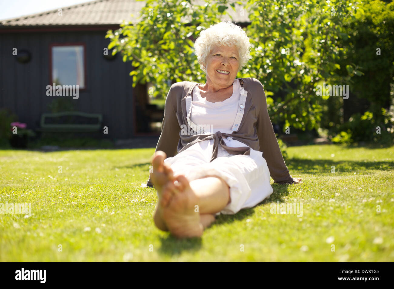 Happy elder woman sitting relaxed on grass in backyard garden and looking at camera smiling - Outdoors Stock Photo