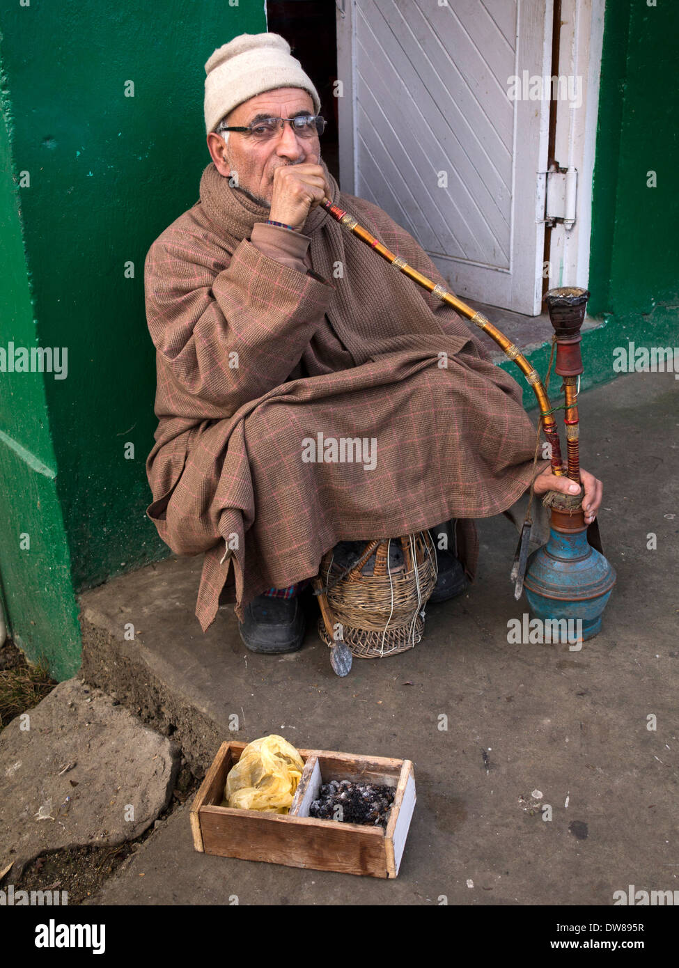 India, Kashmir, Srinagar, man wearing Kashmiri phiren cloak smoking shisha water pipe outside in winter Stock Photo