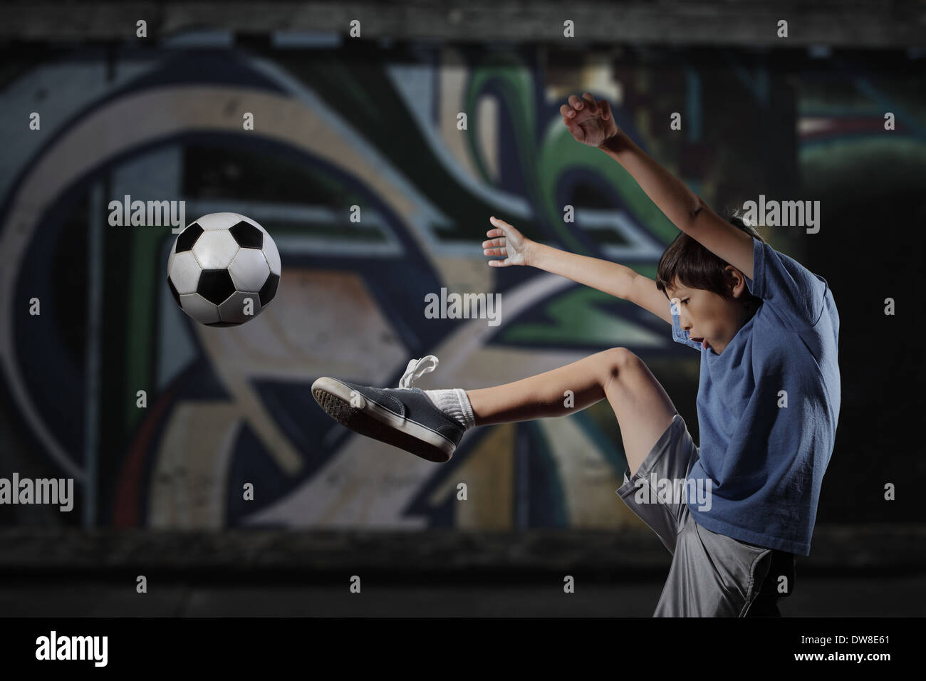 A young boy plays street soccer against a graffiti covered wall - with dramatic lighting and subdued colors Stock Photo