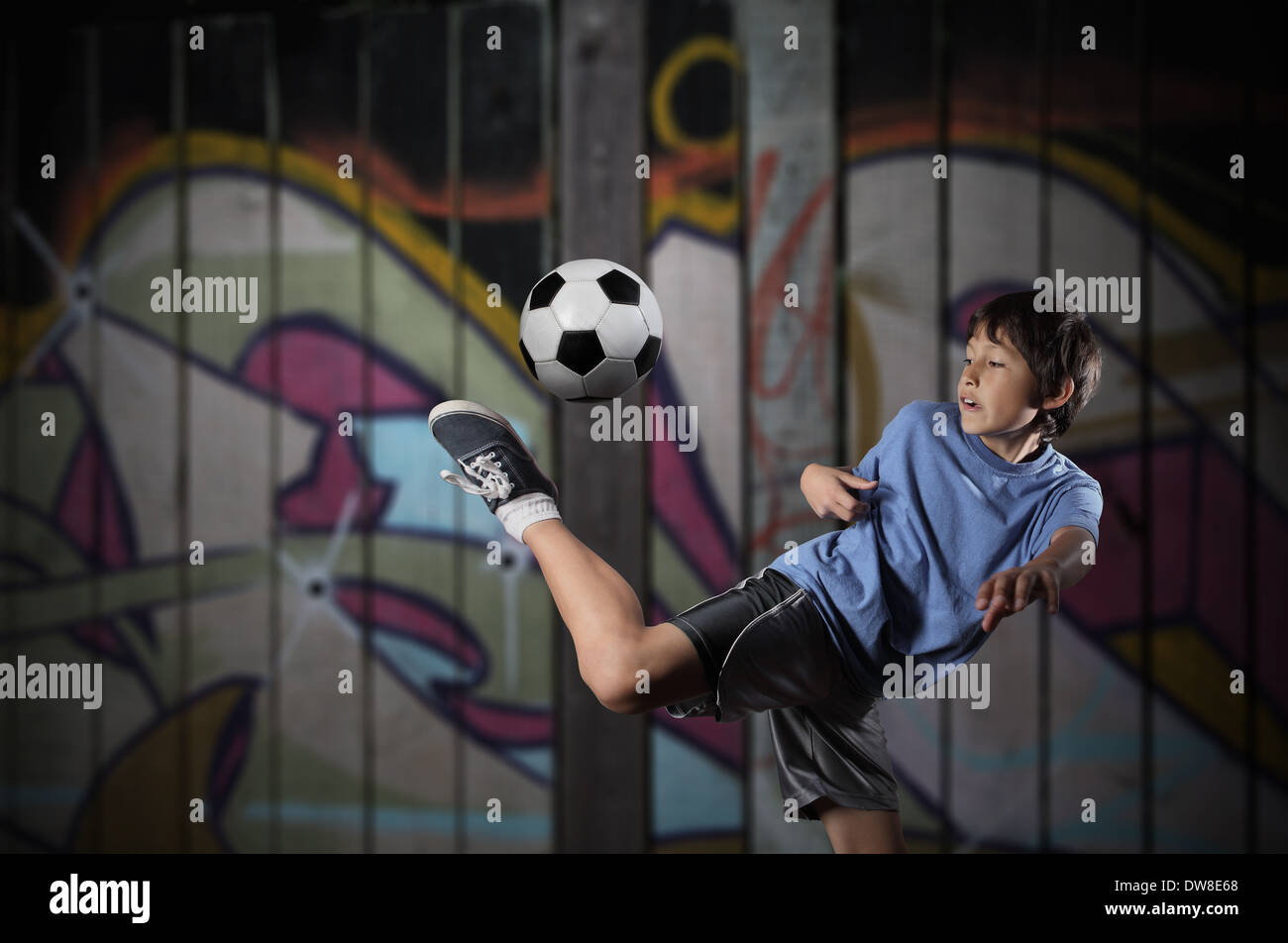 A young boy plays street soccer against a graffiti covered wall - with dramatic lighting and subdued colors Stock Photo