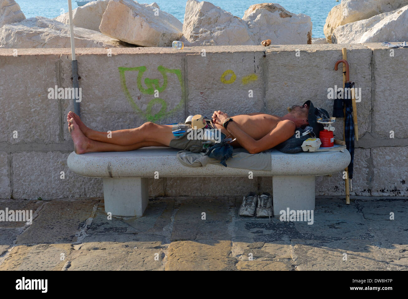 A young male adult sleeping on a stone bench beside all his belongings. Piran. Slovenia Stock Photo