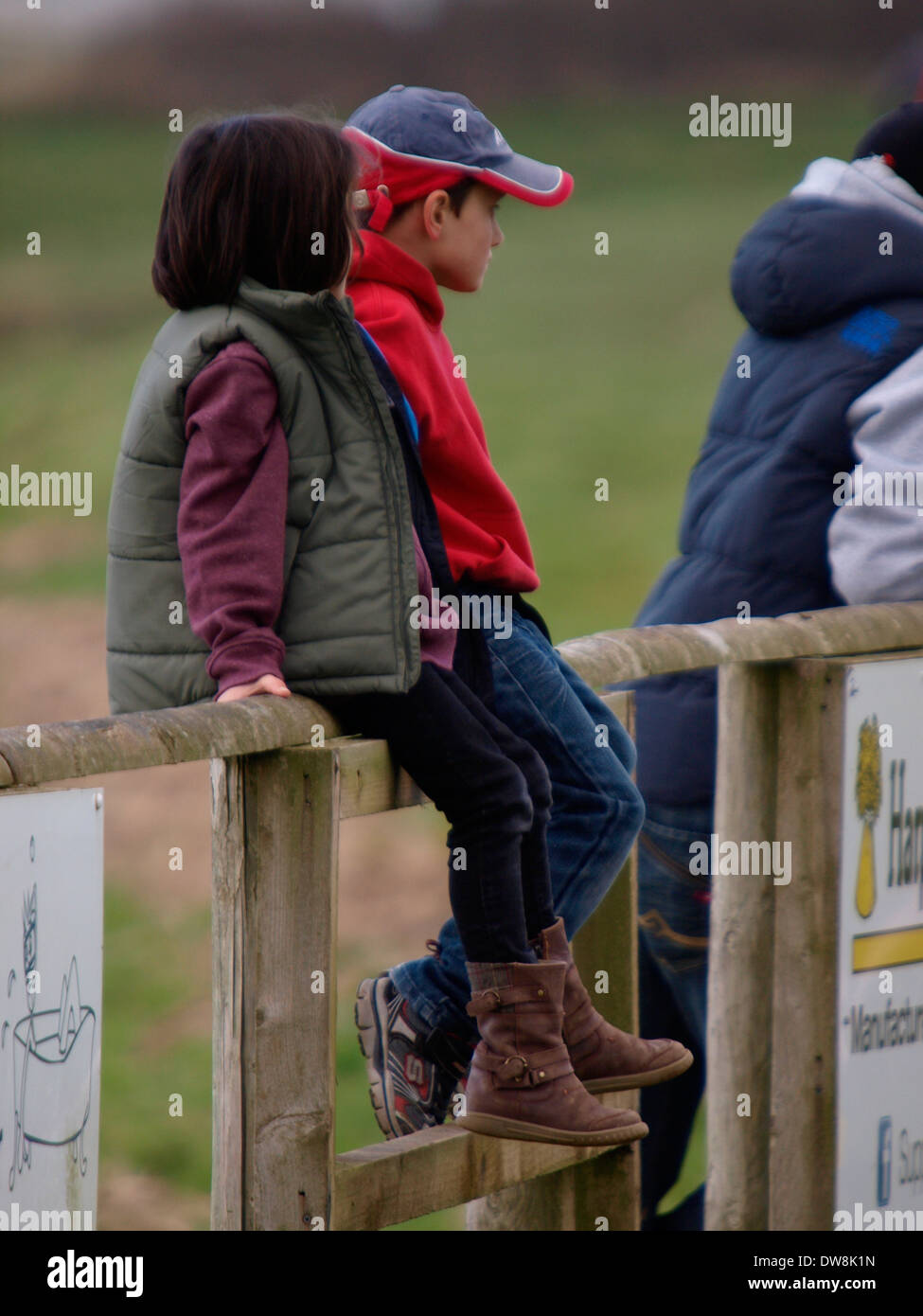 Two children sat on fence watching rugby match, Bude, Cornwall, UK Stock Photo