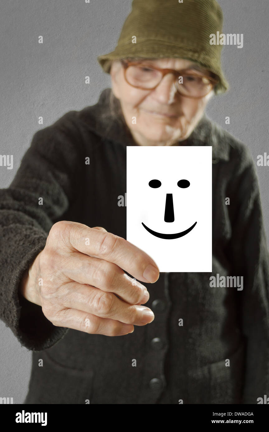 Elderly woman holding card with printed happy emoticon. Selective focus on card and fingers. Stock Photo