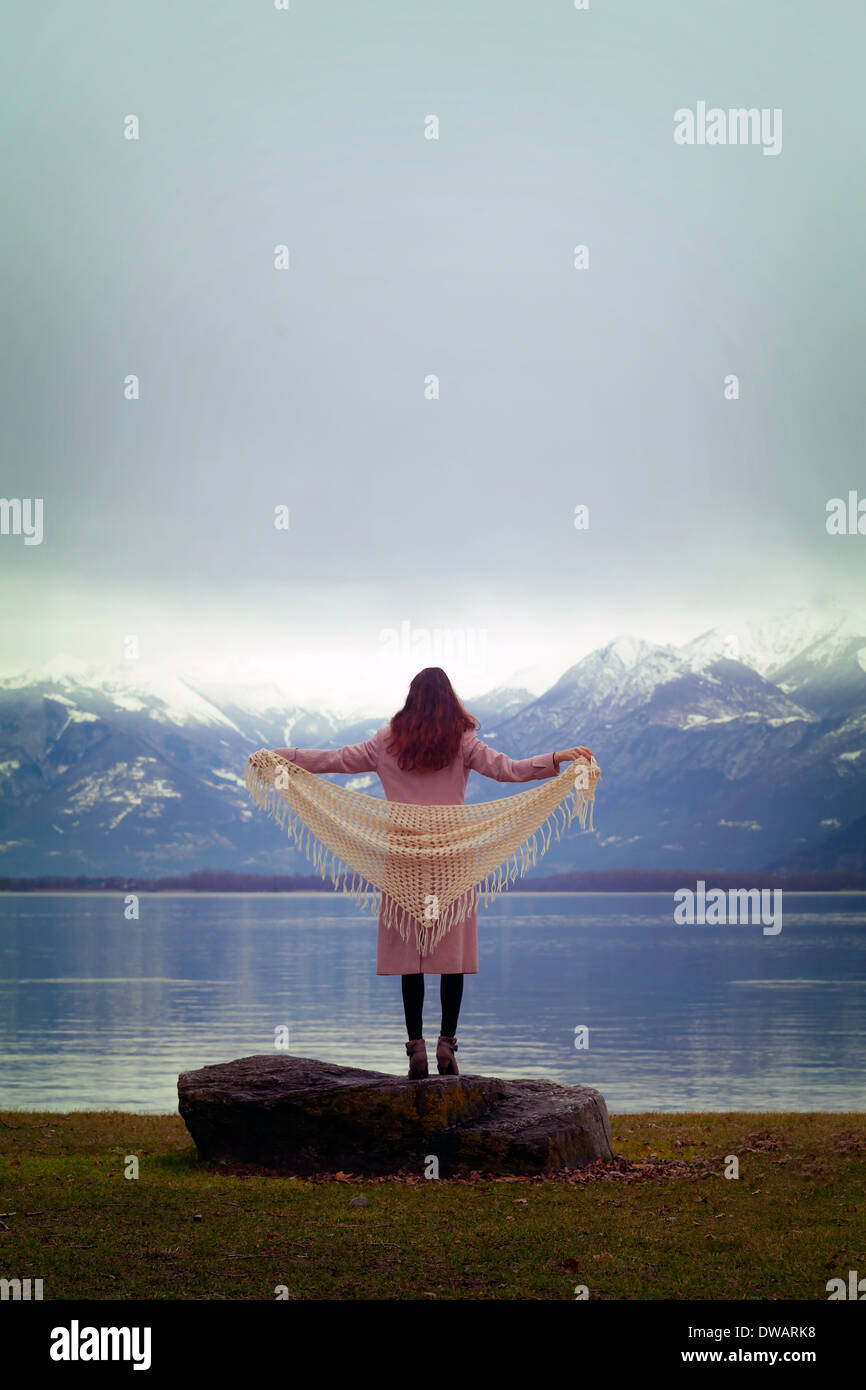 a woman in a pink coat with a white shawl is standing at a lake Stock Photo