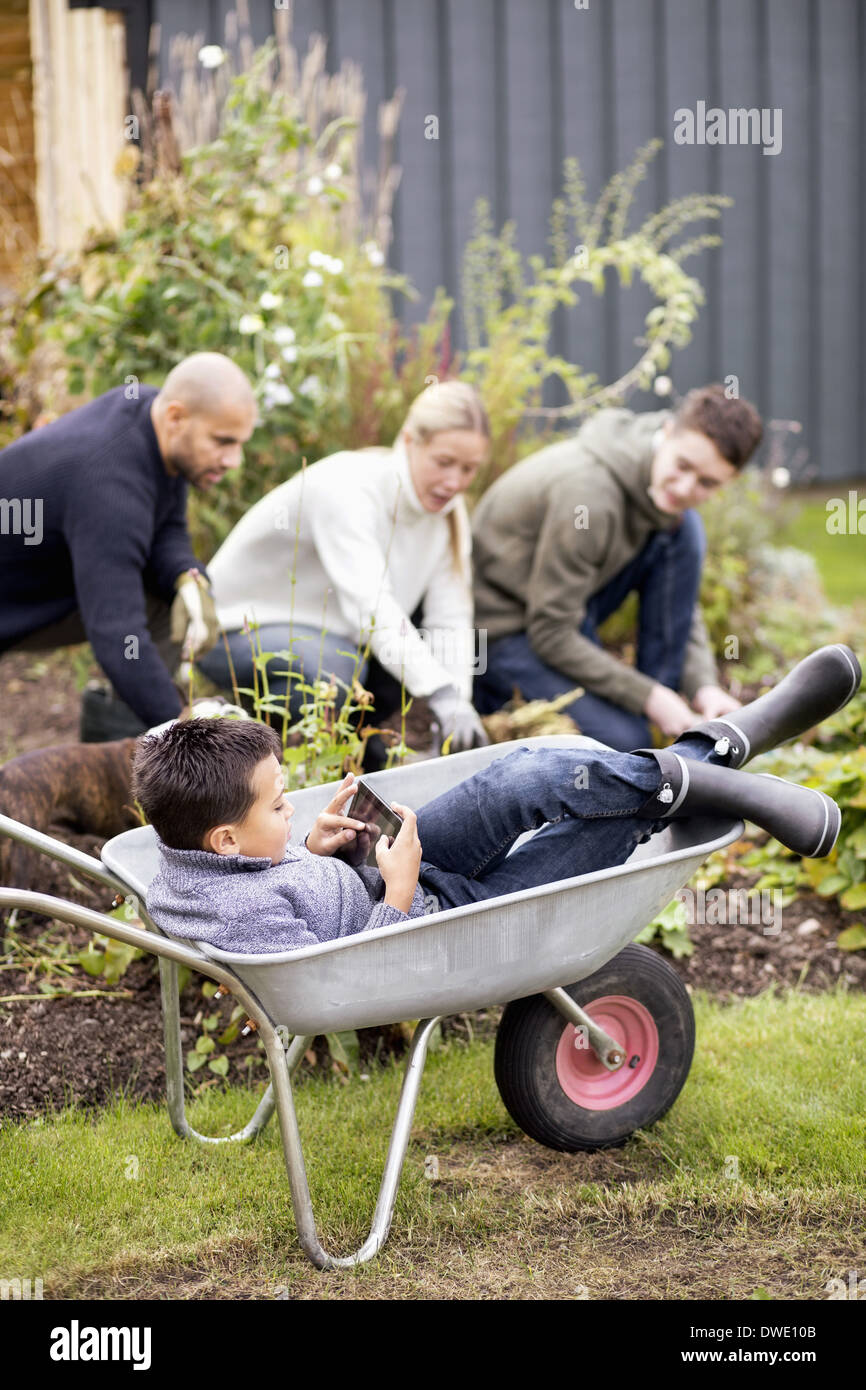 Boy using digital tablet while sitting in wheel barrow with family gardening in background Stock Photo