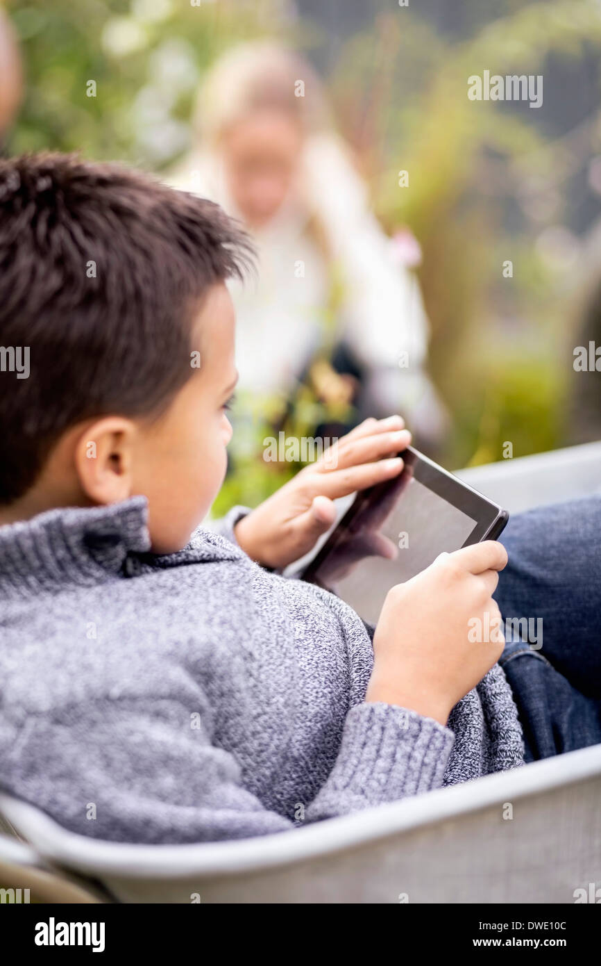 Boy using digital tablet while sitting in wheel barrow with mother gardening in background Stock Photo