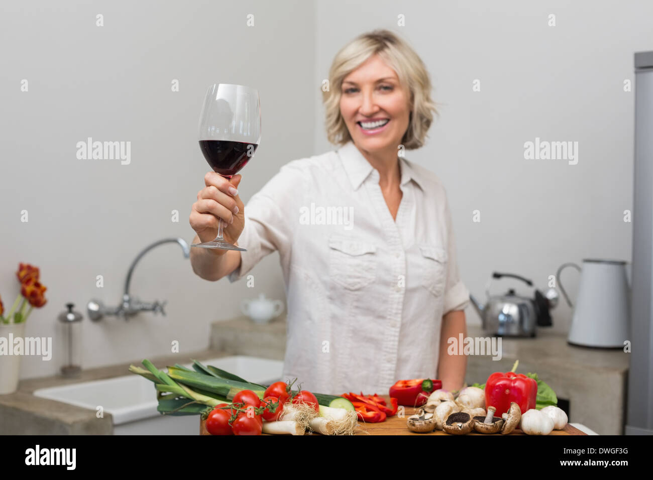 Happy mature woman with vegetables and wine glass in kitchen Stock Photo