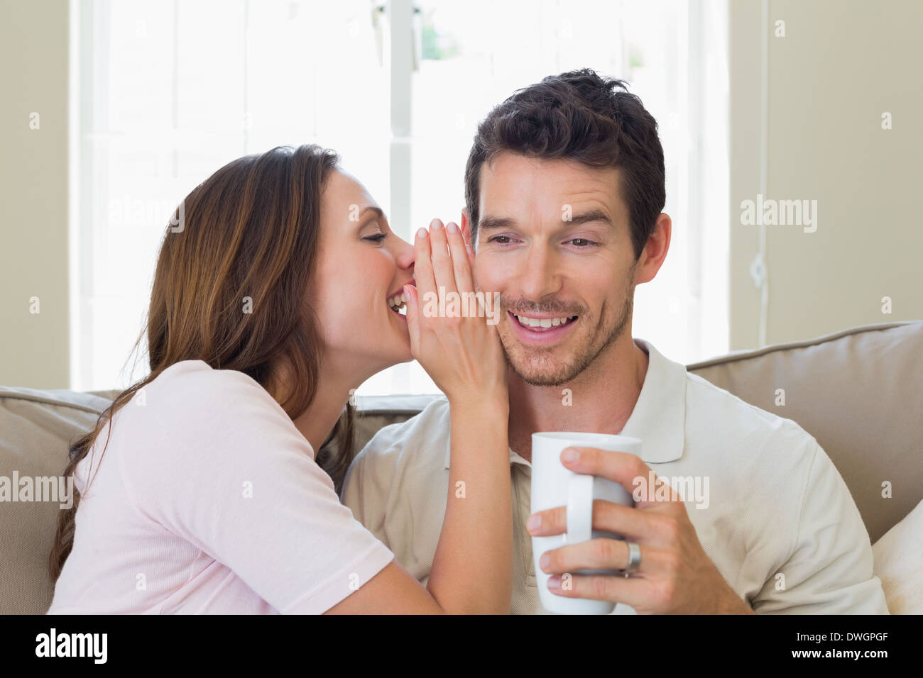 Woman whispering secret into a happy mans ear in living room Stock Photo