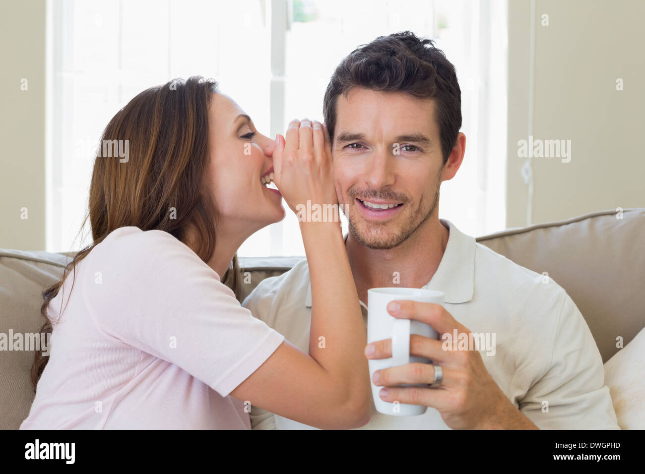Woman whispering secret into a happy mans ear in living room Stock Photo