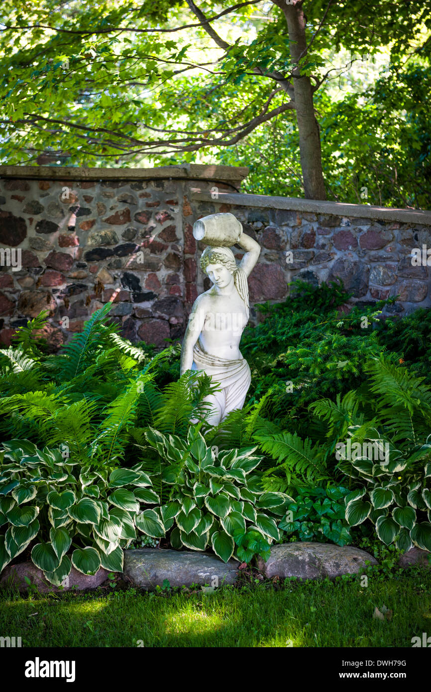 Lush green summer garden with perennial plants and statue near stone wall Stock Photo