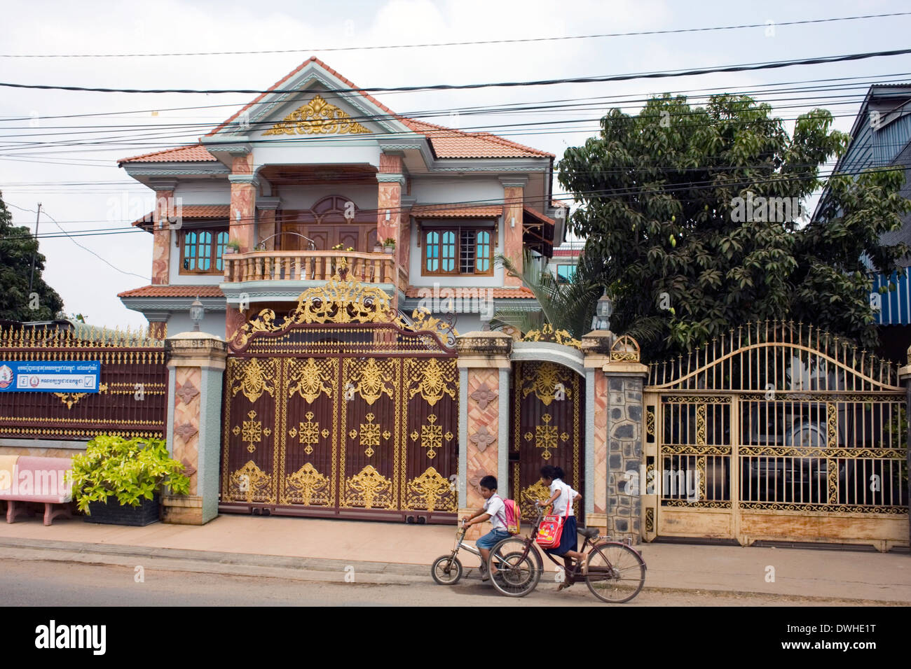 A boy & girl are riding bicycles past a large house belonging to a supporter of Cambodia's ruling CPP in Kampong Cham, Cambodia. Stock Photo