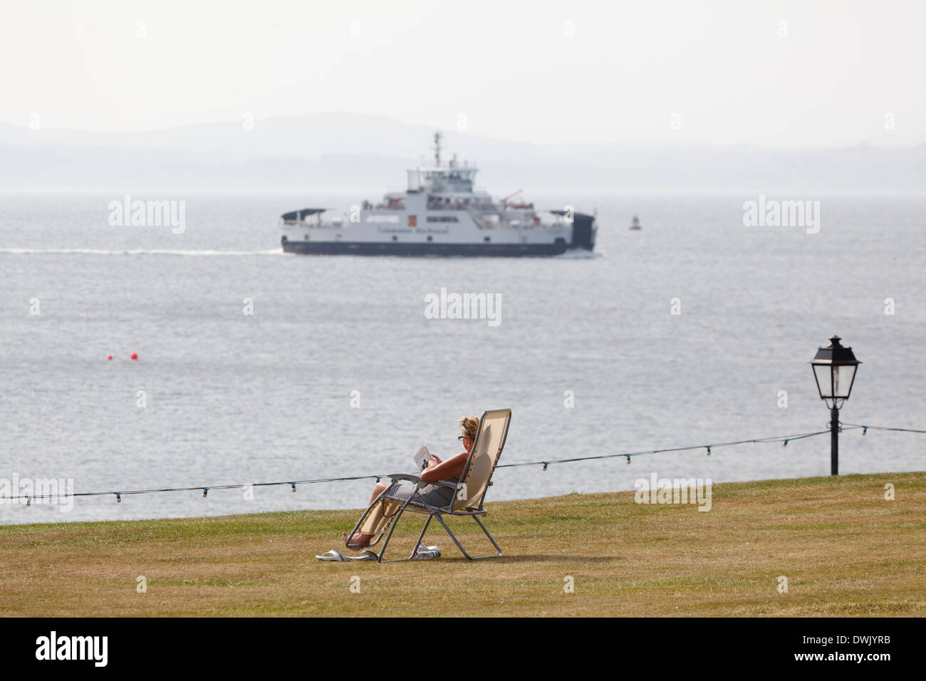 Woman sitting on a deck chair reading a book in the sun beside the Firth of Clyde in the seaside town of Largs, North Ayrshire, Scotland, UK, Europe Stock Photo