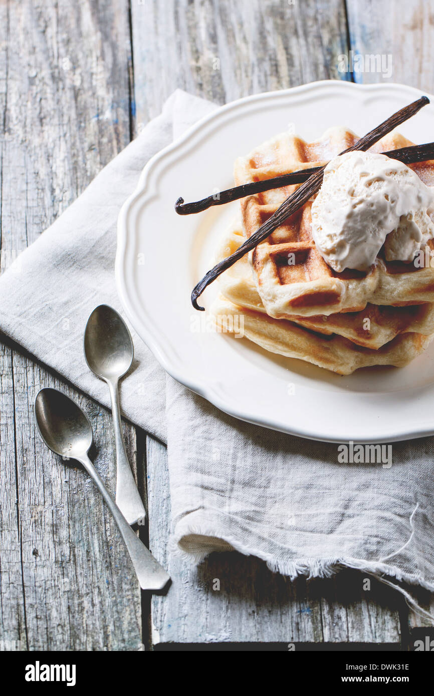 White plate with fresh Belgian waffles, served with ice cream and vanilla sticks over wooden table. Stock Photo