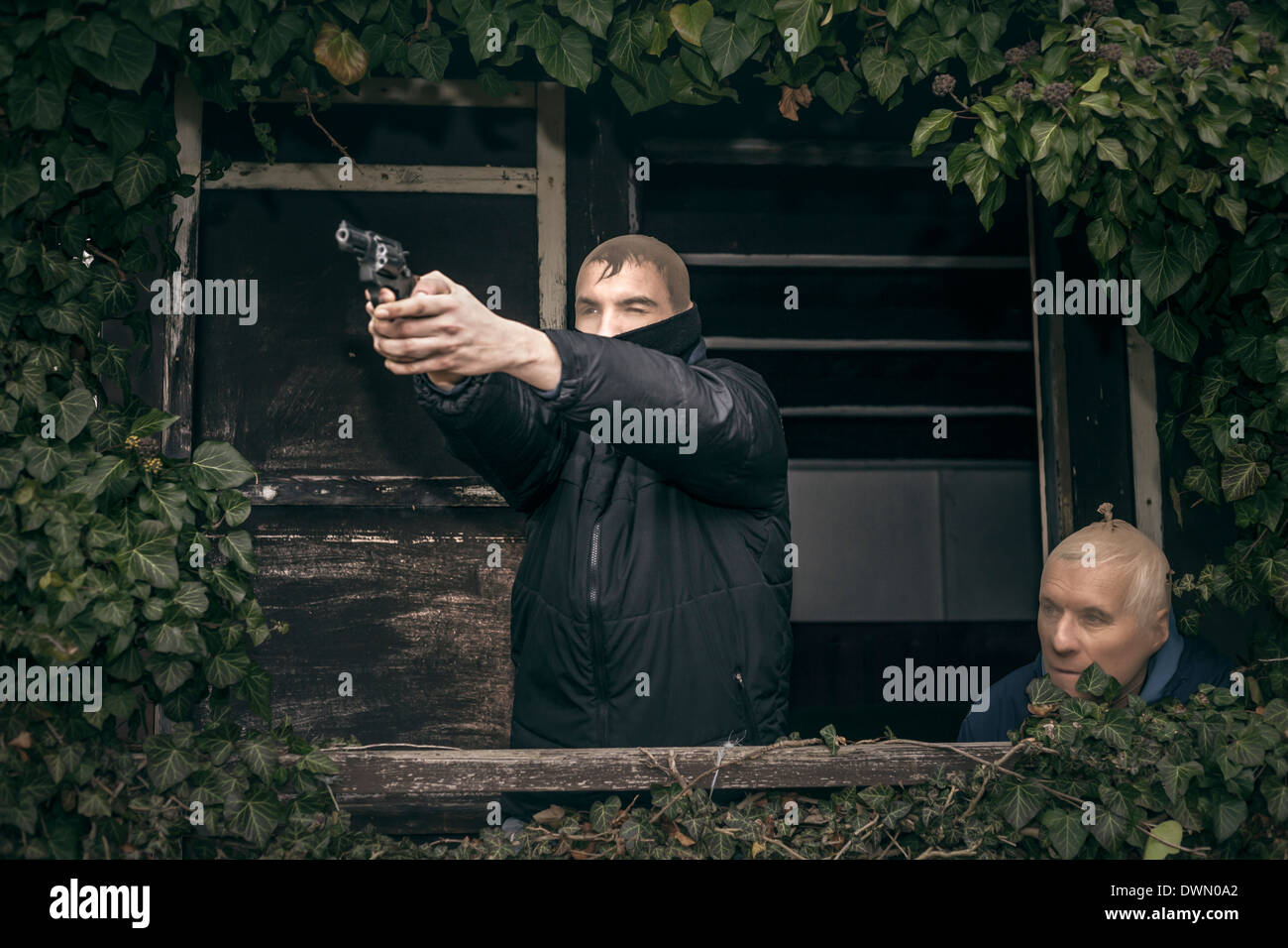 Two masked armed men hiding on overgrown porch of old cabin. Stock Photo