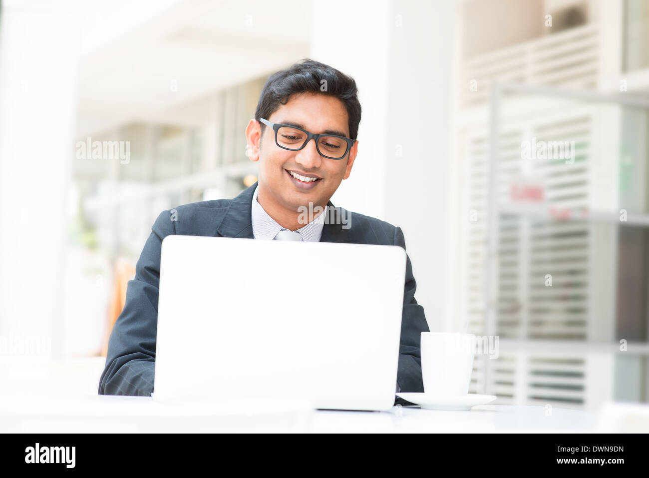 Young Asian Indian businessman using a notebook computer or laptop during office break at cafe, relaxing with a cup of coffee. India male business man, real modern office building as background. Stock Photo