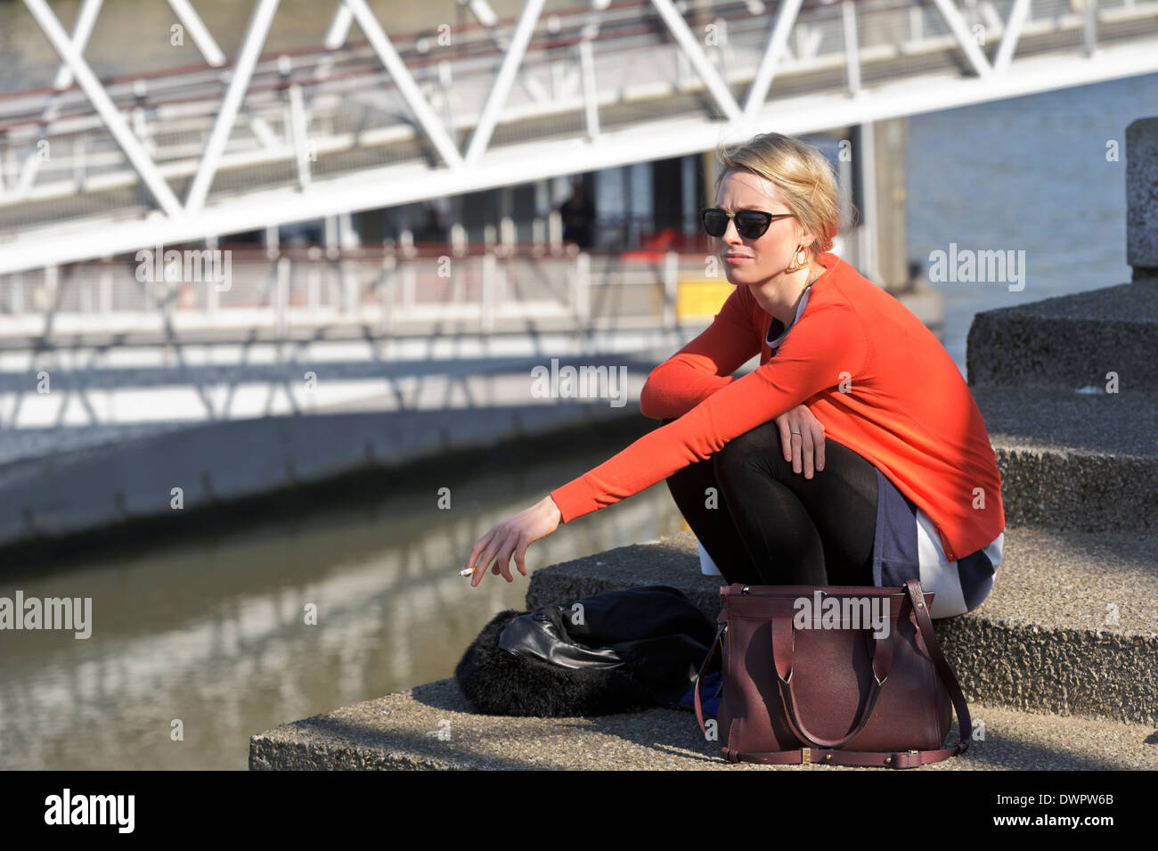 Young blonde woman in red jumper relaxing by the Thames river with a cigarette in her hand, London, England, United Kingdom. Stock Photo