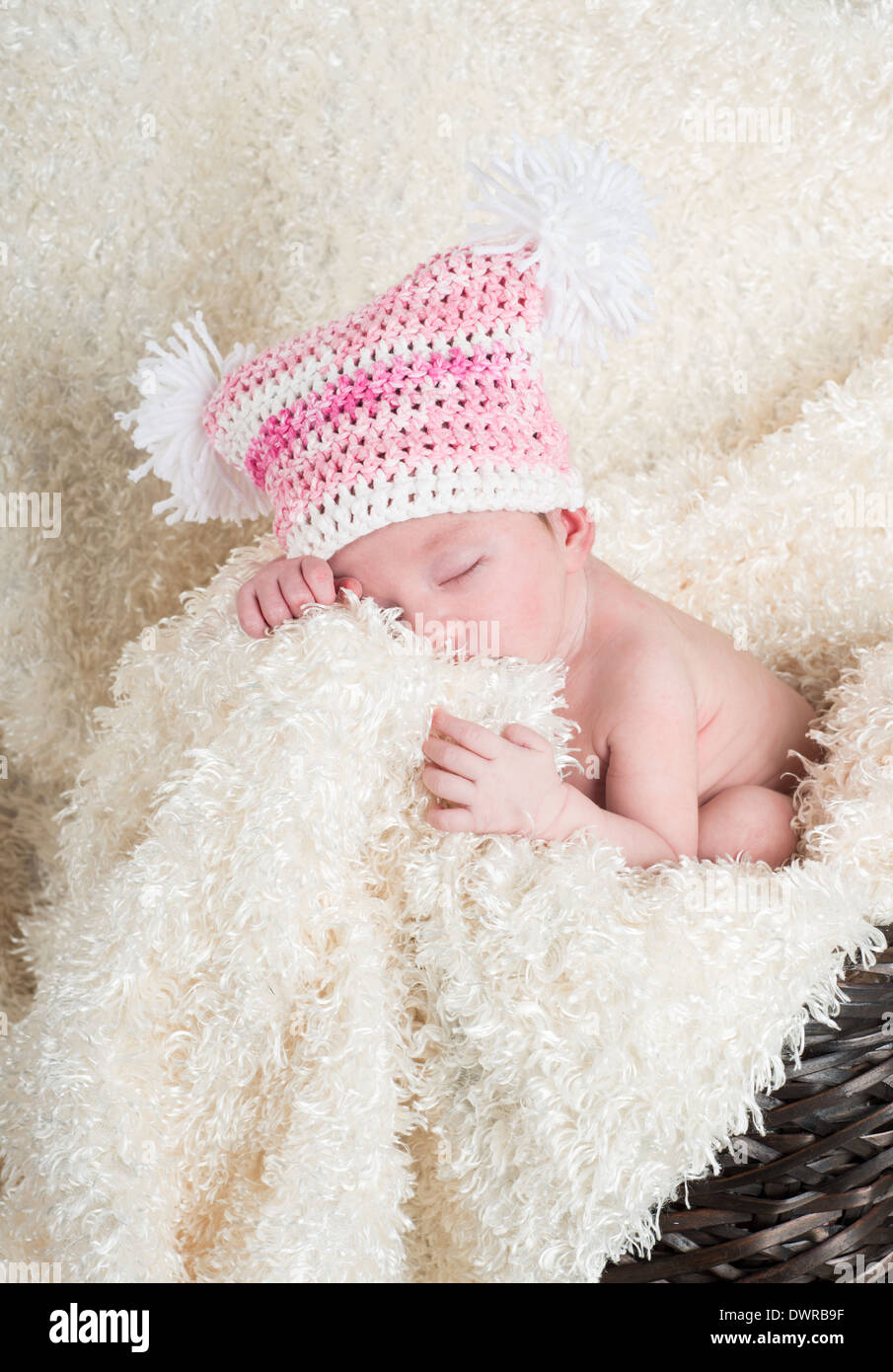 Beautiful newborn baby wearing a pink hat with white pom poms sleeping in a brown woven basket on a soft cream colored blanket Stock Photo