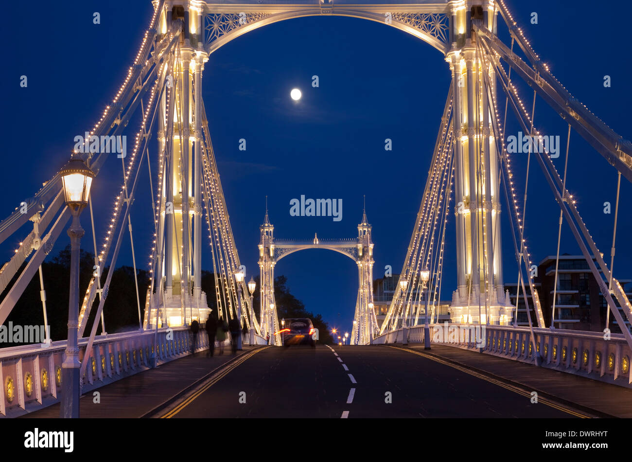 Albert Bridge across the Thames in London, festooned with lights and the moon shining through the night sky behind. Stock Photo