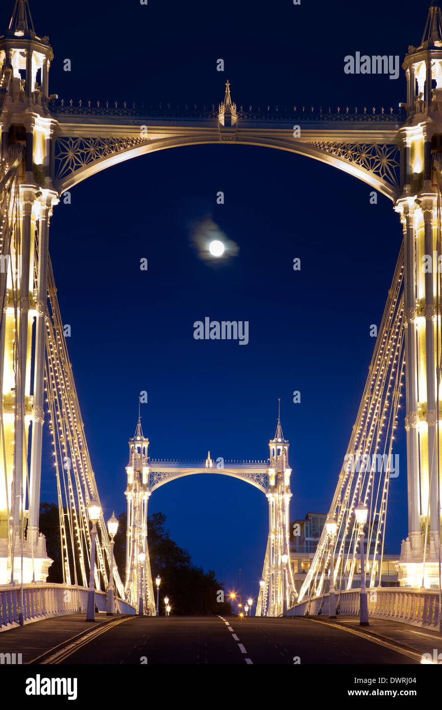 Albert Bridge across the Thames in London, festooned with lights and the moon shining through the night sky behind. Stock Photo