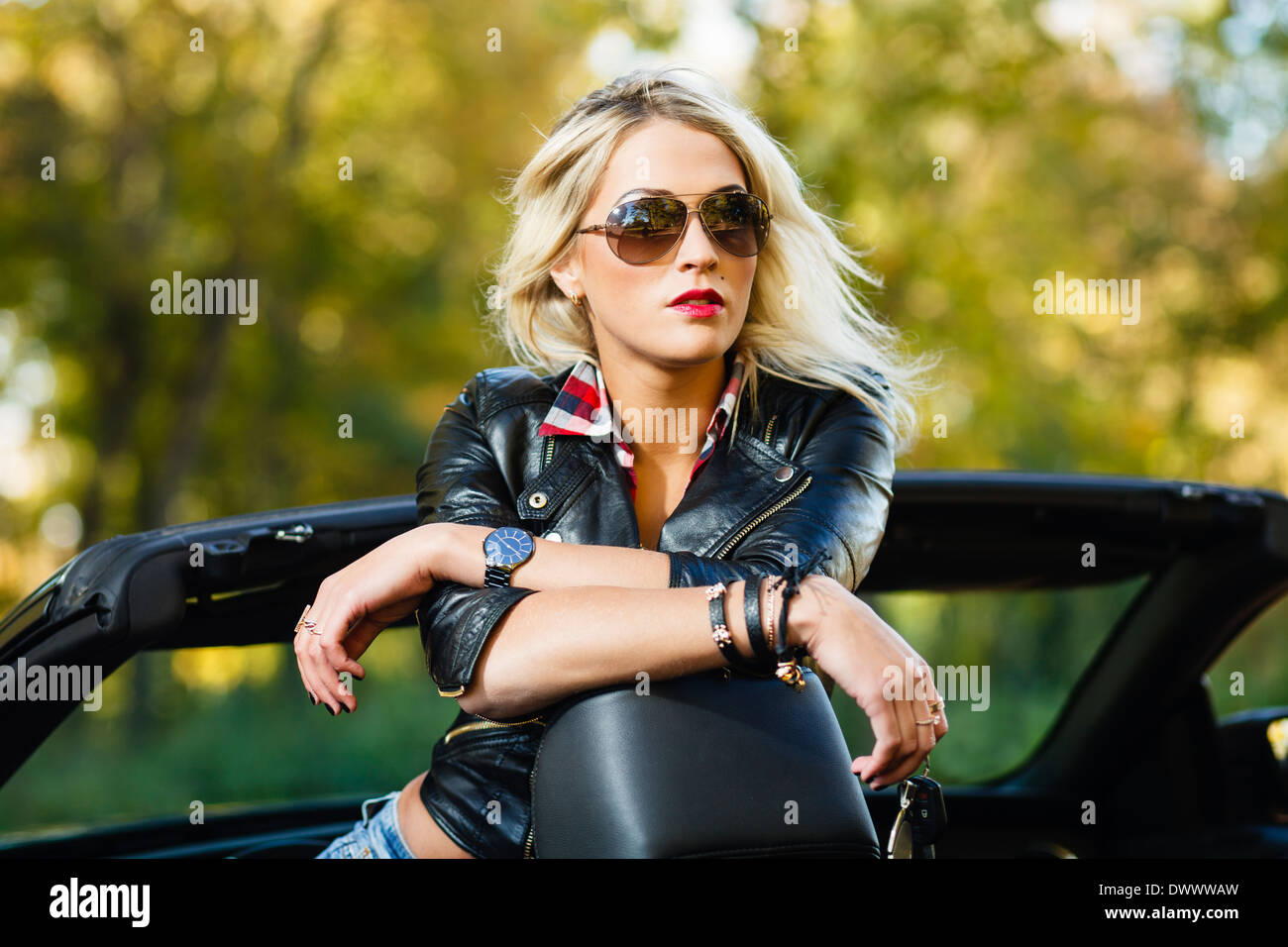 Blond woman in leather jacket and jeans shorts in convertible car. Stock Photo
