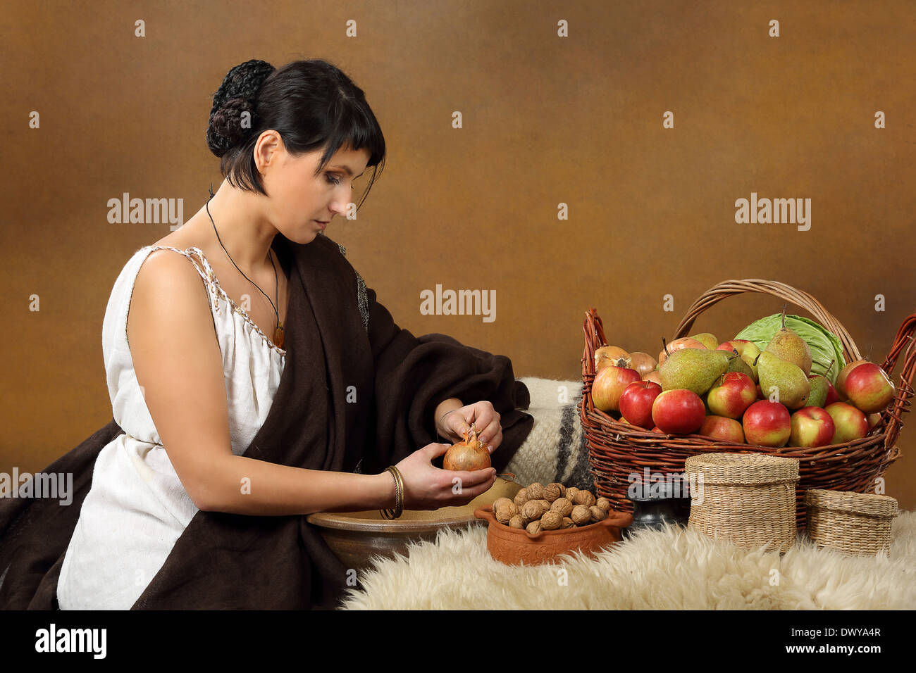 Young Romana peeling the onion with basket full of fruits and vegetables. Concept studio portrait on brown background. Stock Photo