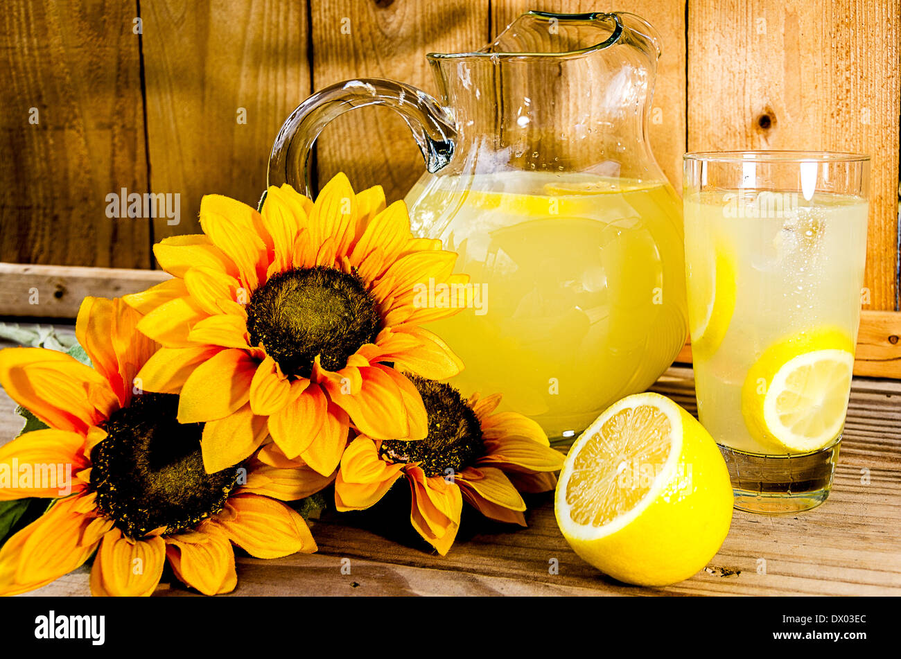 Summer lemonade with lemon slices, pitcher, and sunflowers on wood bench. Stock Photo