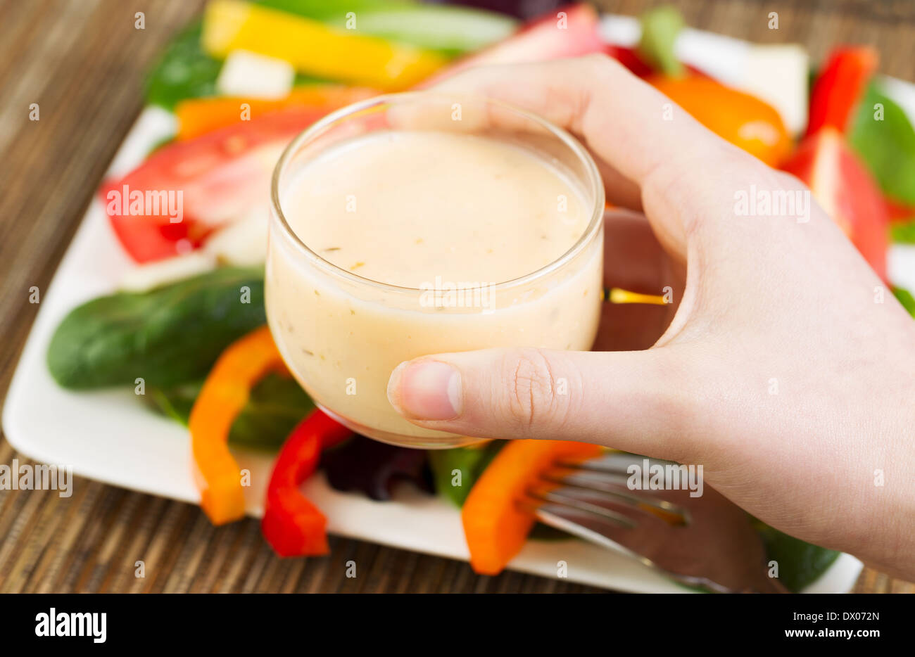 Horizontal photo of female hand holding dressing in small glass bowl with salad and plate in background Stock Photo
