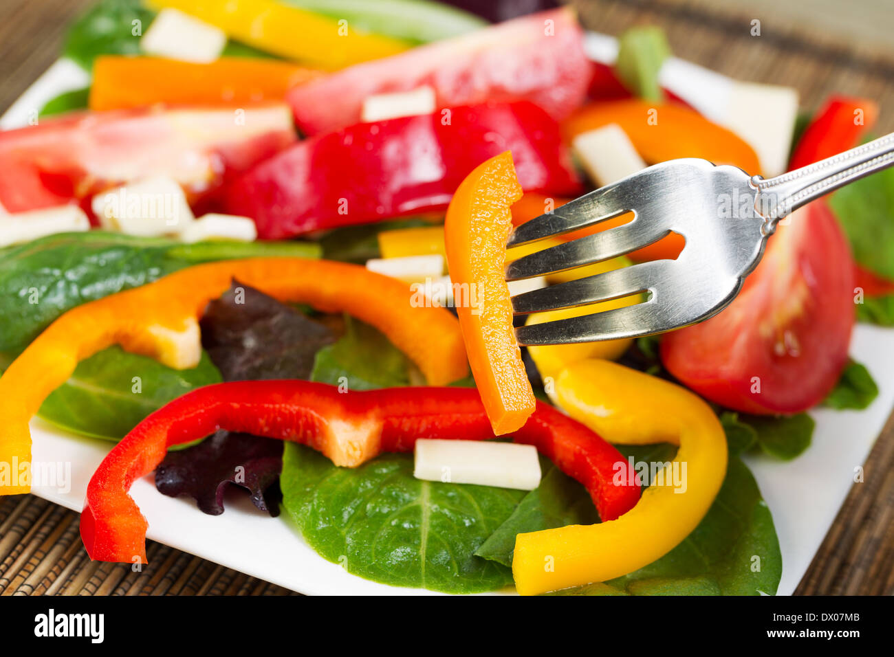 Horizontal photo of a fork holding a single piece of yellow bell pepper with salad and plate in background Stock Photo