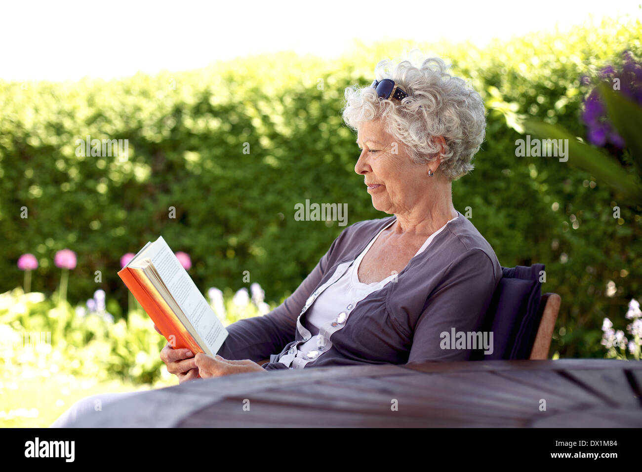 Image of relaxed elder woman sitting in her backyard reading a book - Senior lady reading a novel in backyard garden Stock Photo