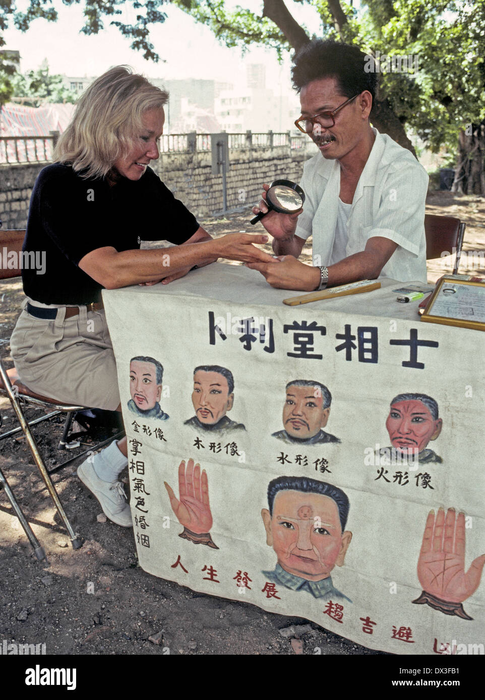 Outdoors under the shade of trees, a Chinese palm reader studies the hand of an American woman who is touring Macau in the People's Republic of China. Stock Photo
