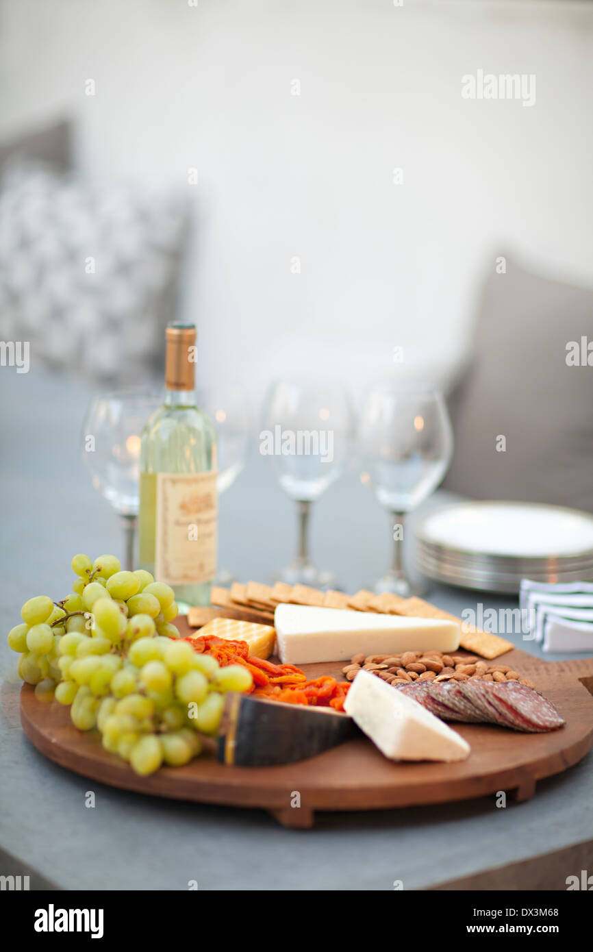 Cheese, crackers, nuts, grapes and salami on wooden cheeseboard next to white wine, tilt Stock Photo