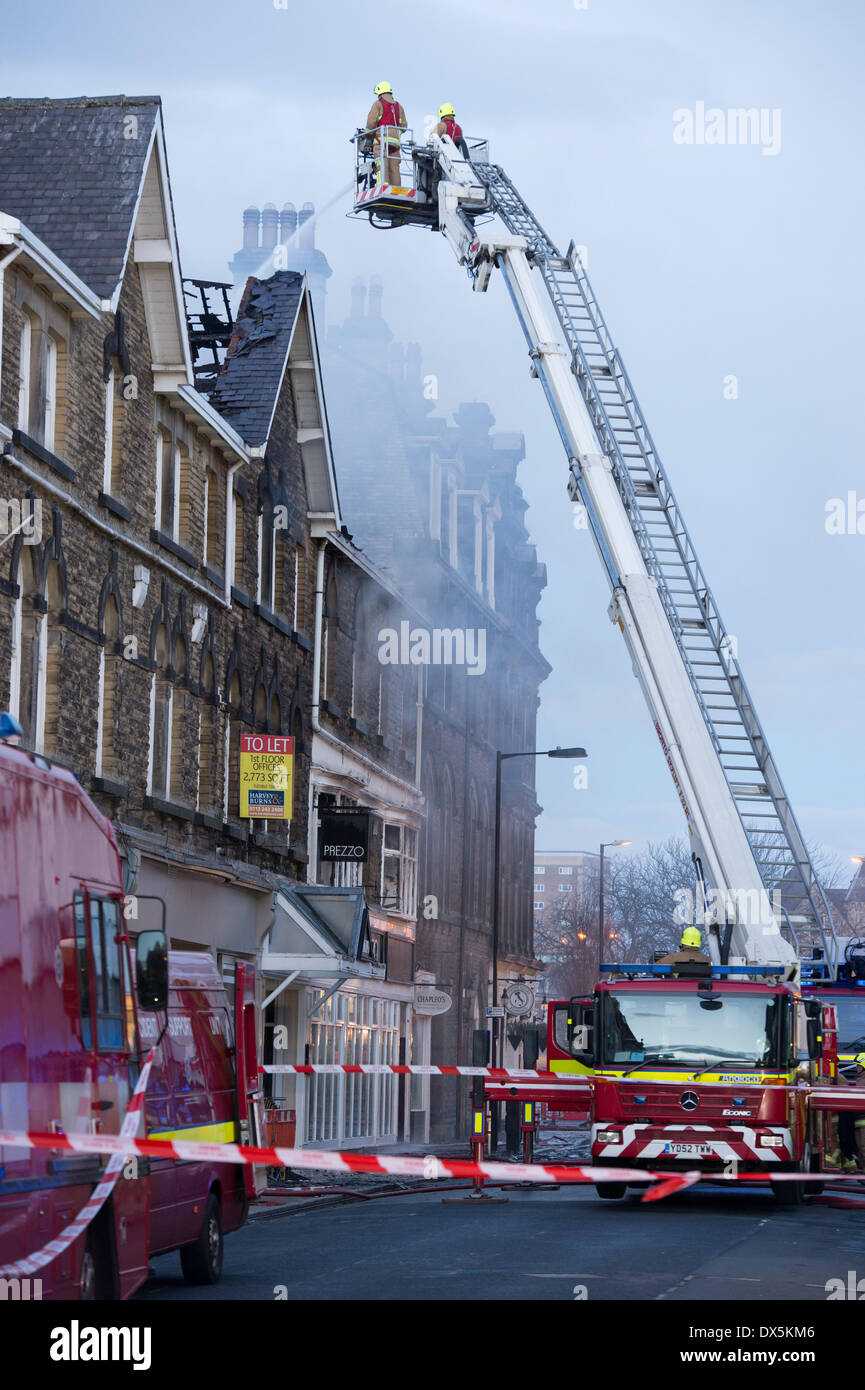 Brave firefighter crew up high ladder (from engine) tackle fire with water hose, at town centre building - Harrogate, North Yorkshire, England, UK. Stock Photo