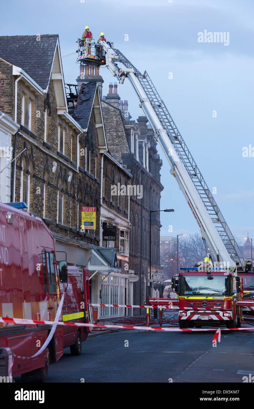 Brave firefighter crew up high ladder (from engine) tackle fire with water hose, at town centre building - Harrogate, North Yorkshire, England, UK. Stock Photo