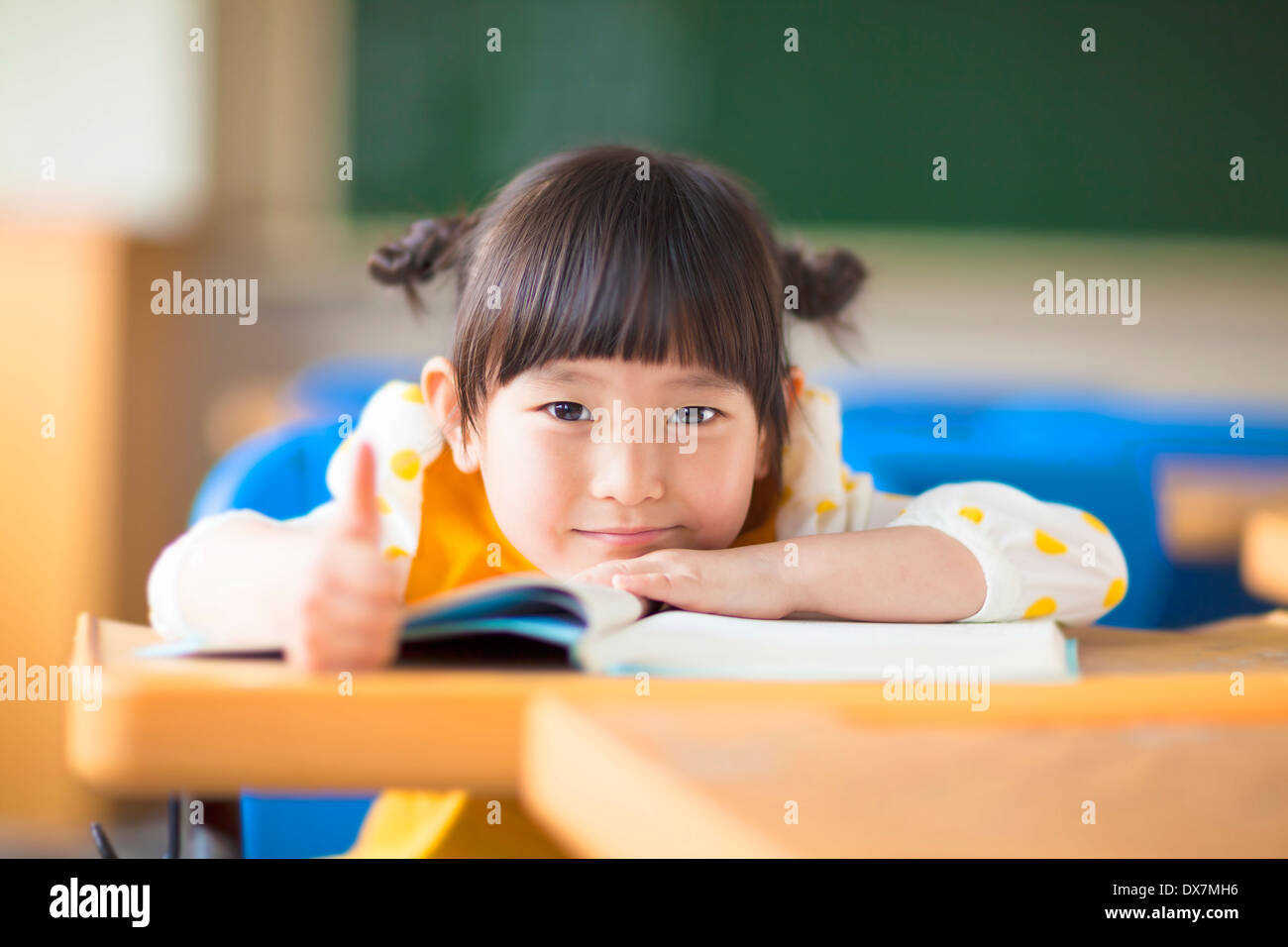smiling kid lie prone on a desk and thumb up in a classroom Stock Photo