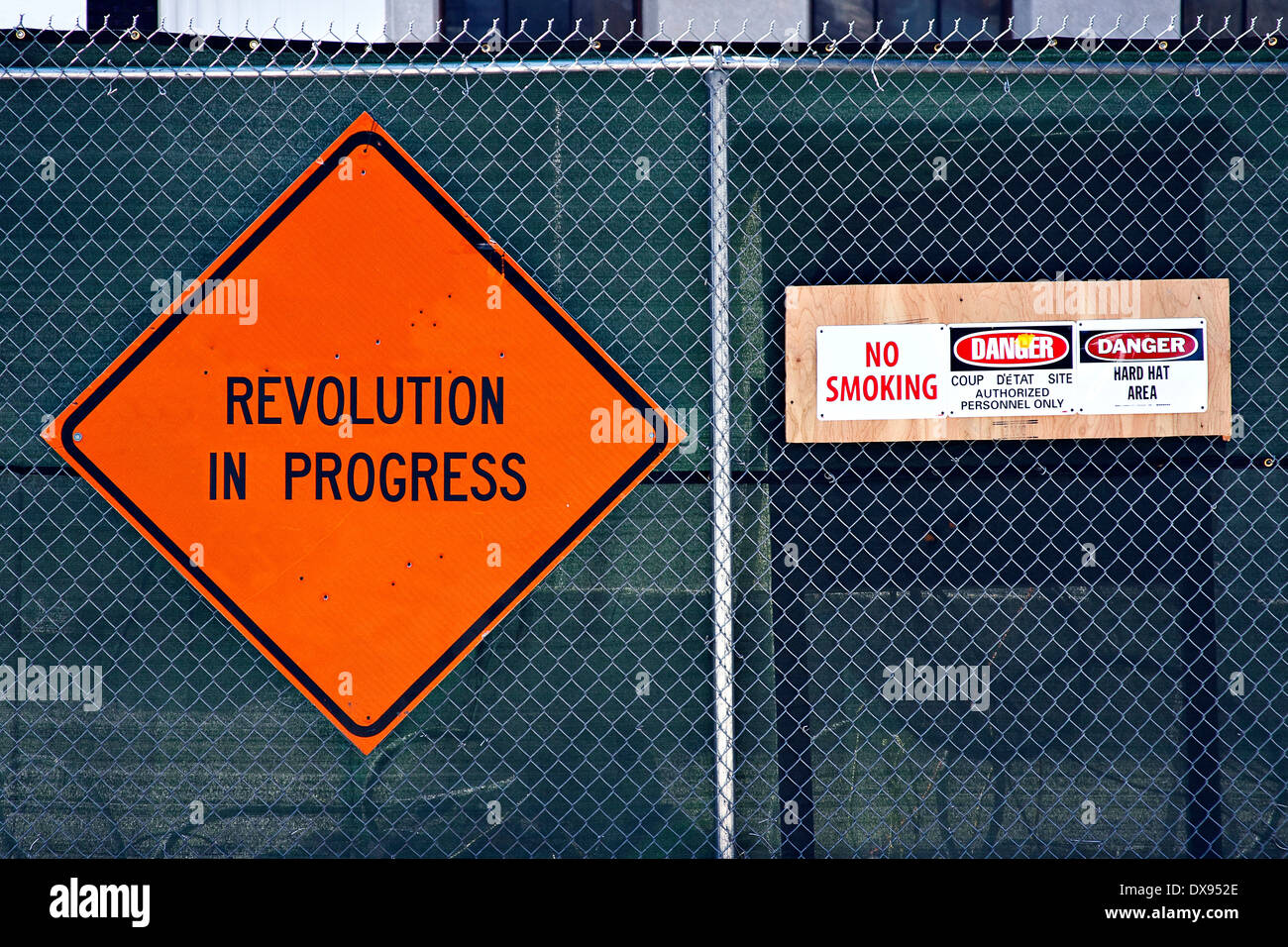 Orange 'REVOLUTION IN PROGRESS' sign on chainlink fencing along with 'DANGER coup d'état site' stapled on wooden board on right Stock Photo
