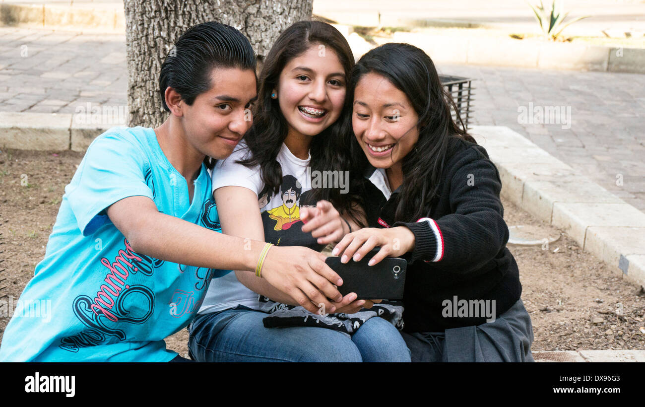3 good looking high school kids take a cellphone selfie while sitting on tree pocket curb in historic center district Oaxaca Stock Photo