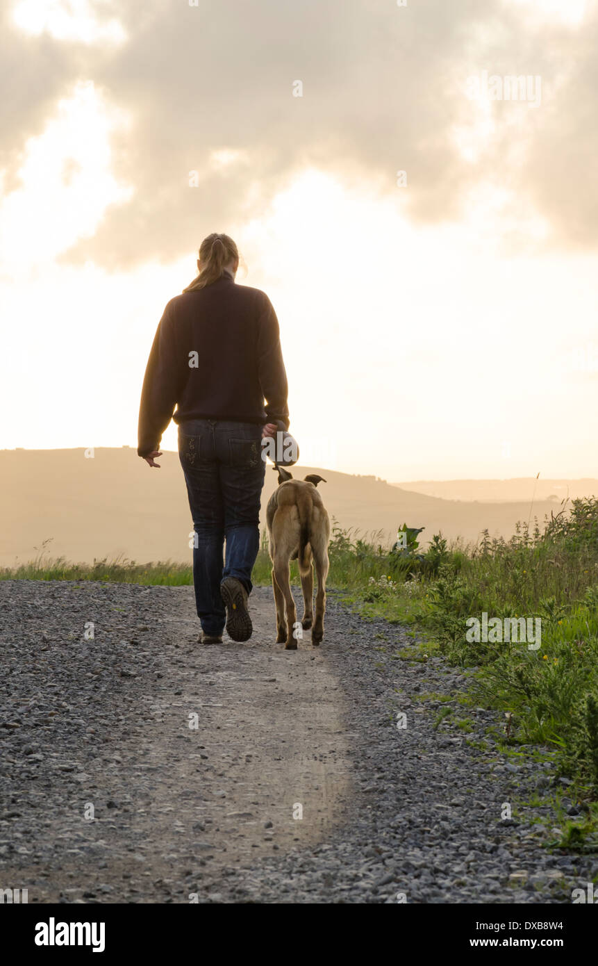 A young woman walking a dog on a stony path, moving away from the camera into the setting sun. Stock Photo
