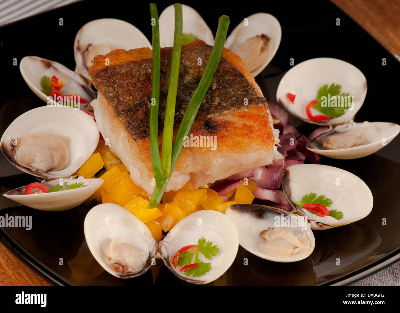 Crisp seared cod fillet on a bed of chives garnished with almejas, clams, and chili on a black plate Stock Photo