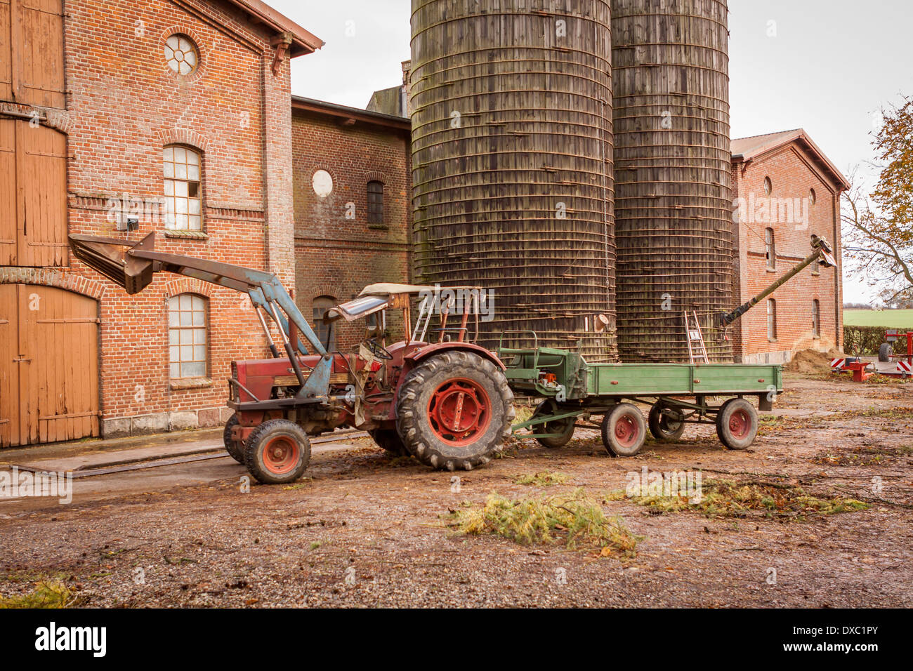Tractor and trailer parked in a farmyard in front of a large multistorey barn and cylindrical silos with a famer just exiting the tractor after coming in from the fields Stock Photo