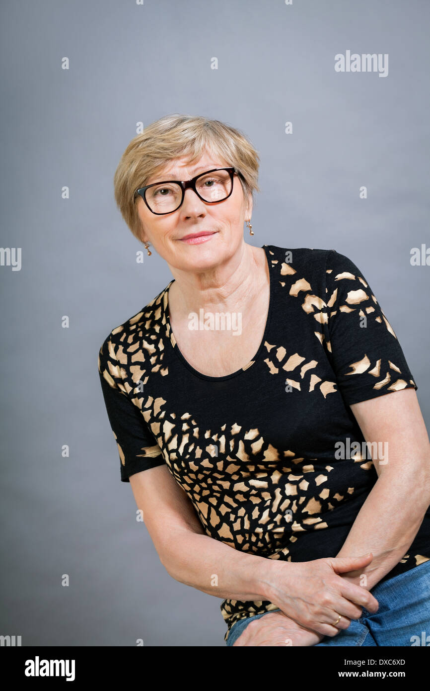 Attractive blond senior woman wearing glasses sitting with clasped hands in a relaxed position smiling at the camera, on grey Stock Photo