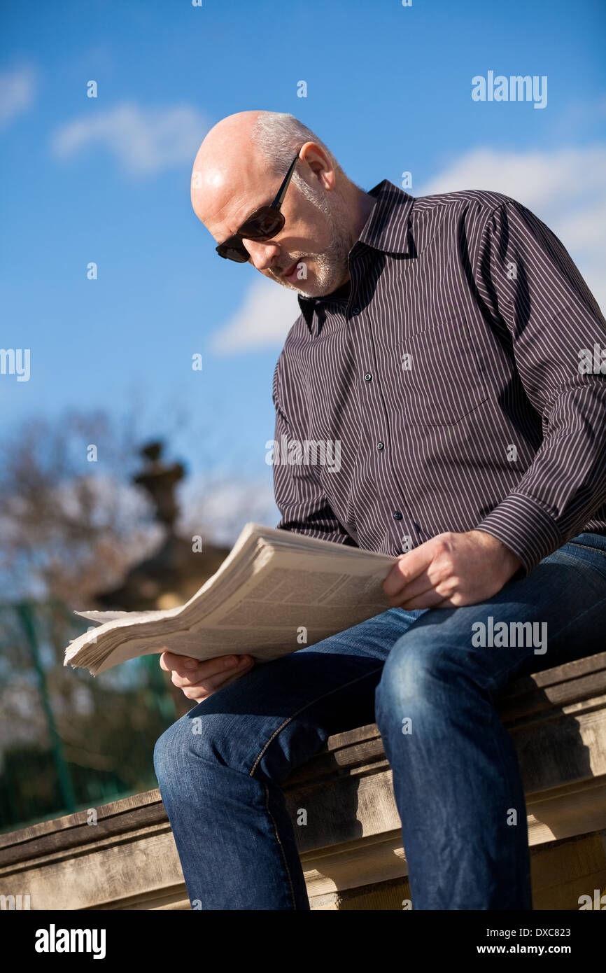 Middle-aged bald man sitting in the sunshine reading a newspaper on a stone wall in an urban environment with his jacket folded up alongside him Stock Photo