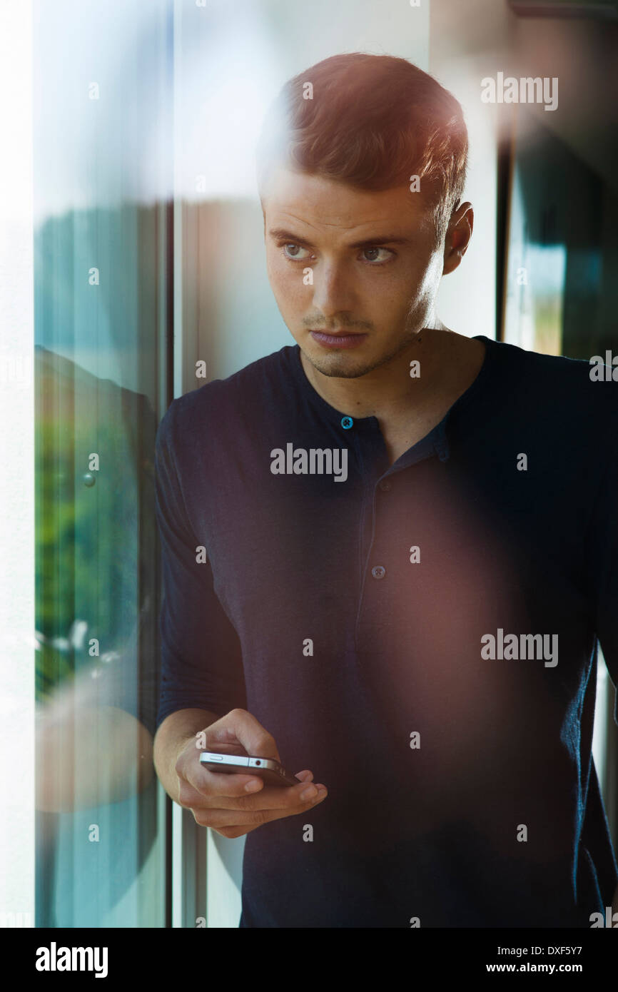 Young Man with Cell Phone Looking out Window, Mannehim, Baden-Wurttemberg, Germany Stock Photo