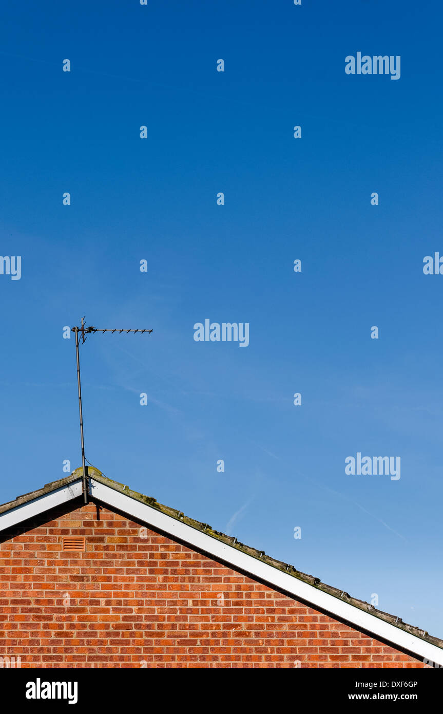 TV antenna / aerial at apex of sloping tiled roof on top of red brick wall against mainly blue sky with hazy clouds. Stock Photo