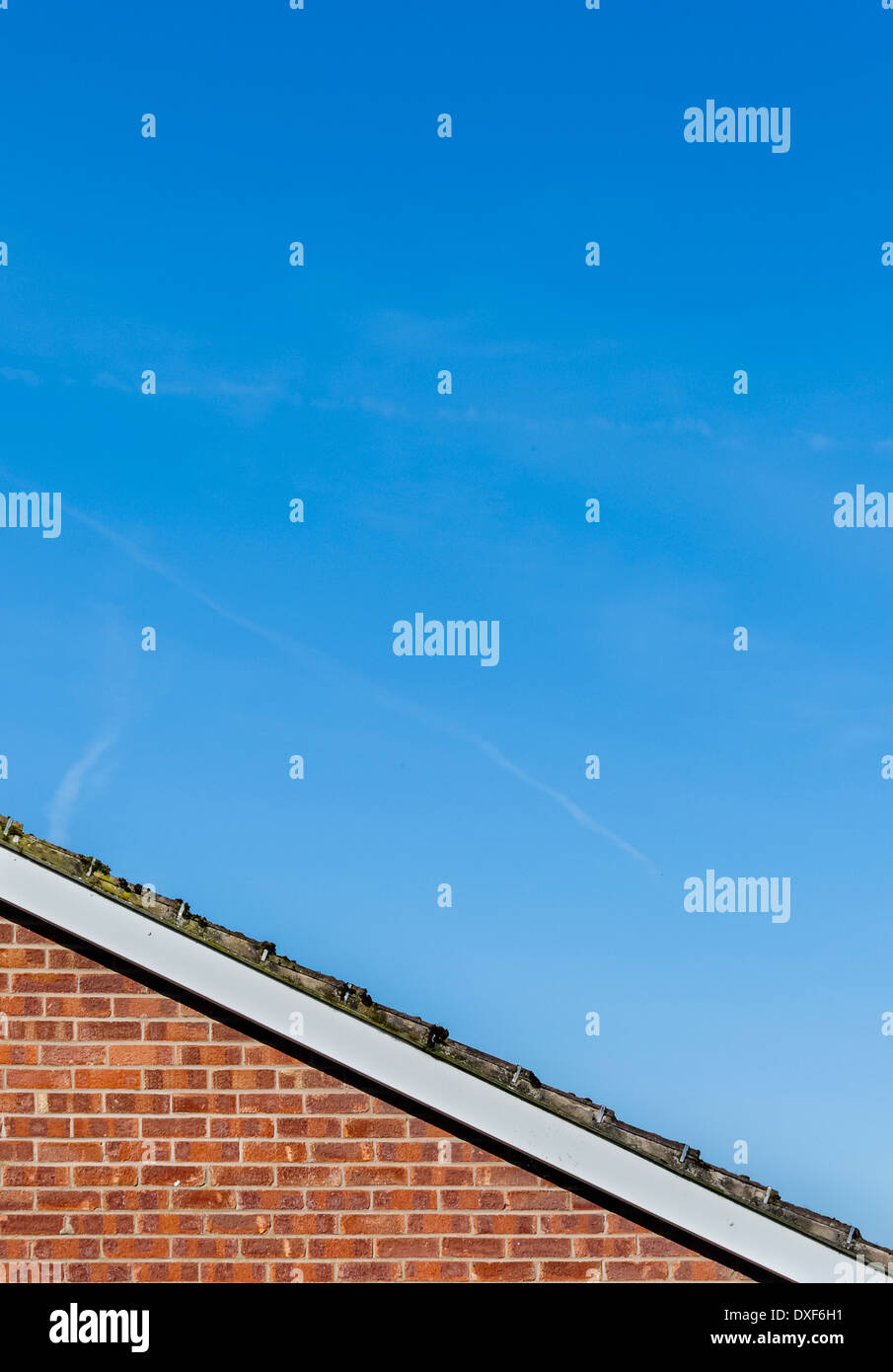 Edge of sloping tiled roof on top of red brick wall against mainly blue sky with hazy clouds. Stock Photo