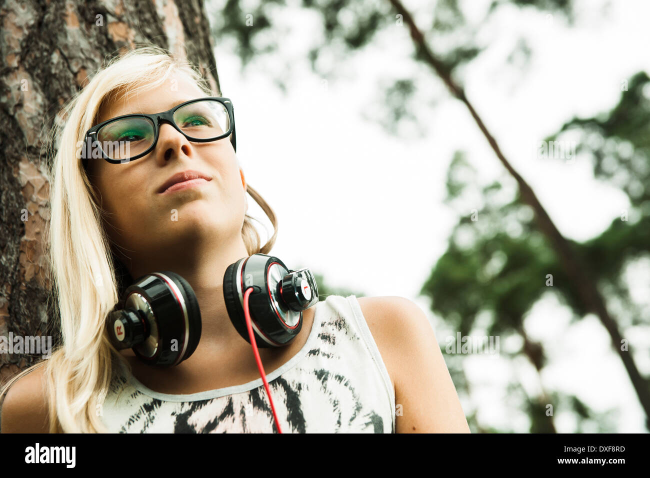Portrait of girl wearing eyeglasses, standing next to tree in park, with headphones around neck, looking upward, Germany Stock Photo