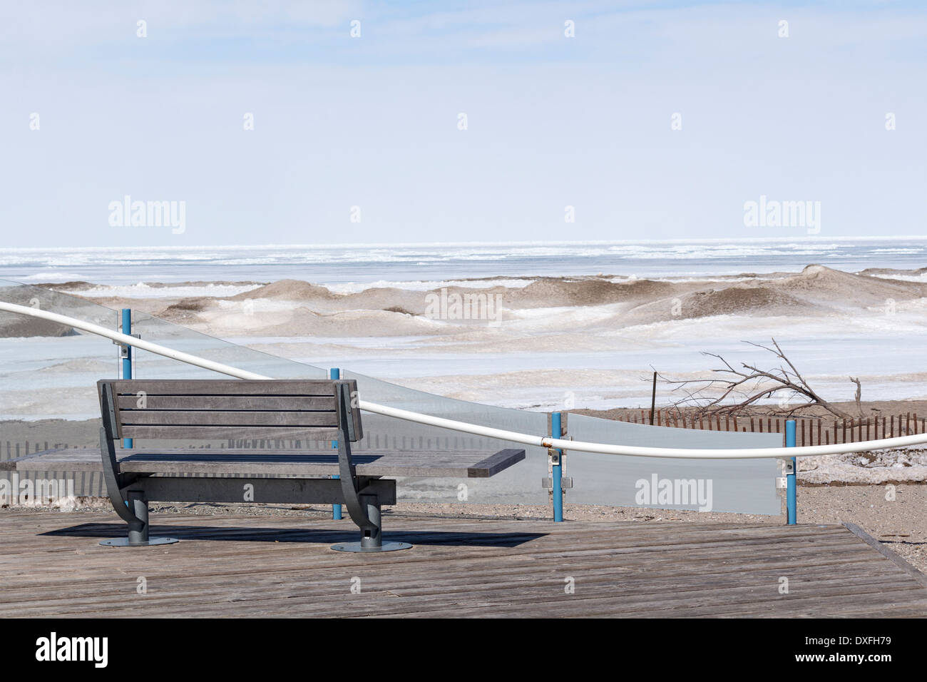 An empty bench looks over a frozen Lake Huron with sand and snow mixed drifts on the lake. Stock Photo