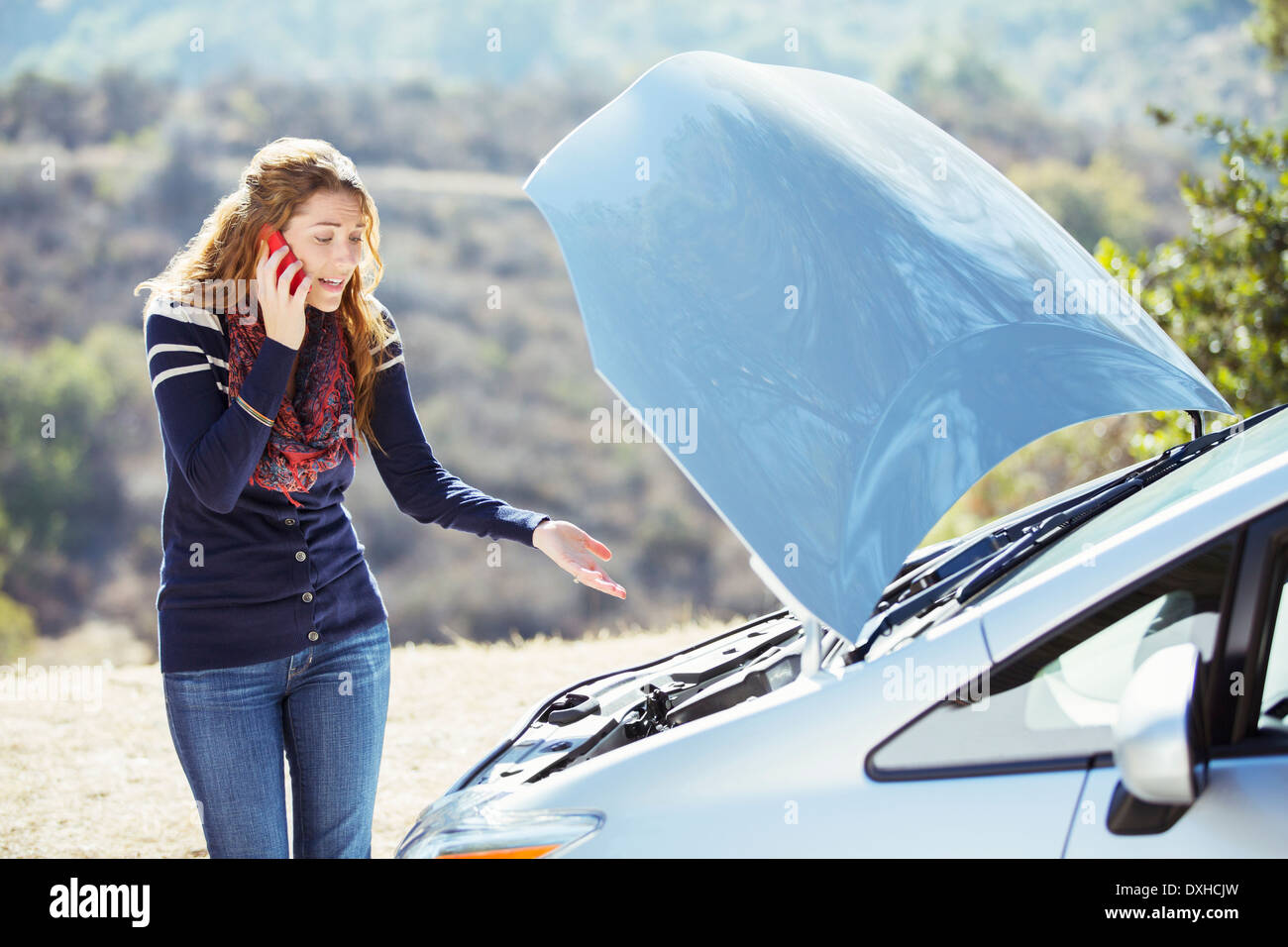 Frustrated woman talking on cell phone with automobile hood raised at roadside Stock Photo